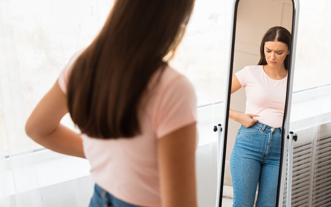 Unhappy woman gaining weight touching belly standing at mirror indoors